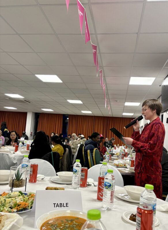 A woman in red clothing stands with a microphone, speaking to people sitting at round tables. Food is laid out on the tables.