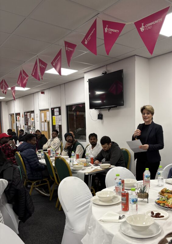 A woman standing with a microphone in front of a group of people sitting at tables.